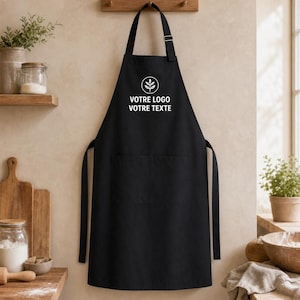 Puede incluir: Un delantal negro que presenta un icono circular blanco con un gráfico de planta y el texto VOTRE LOGO VOTRE TEXTE impreso en blanco en el pecho. El delantal incluye una correa para el cuello ajustable con hebilla metálica y un bolsillo frontal.