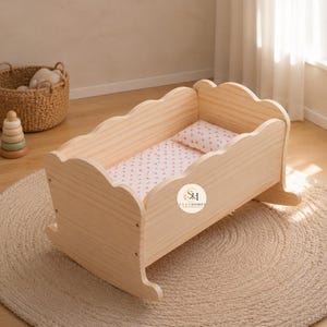 May include: A light-colored wooden doll cradle with a scalloped edge design. The cradle features a small rectangular pillow and a mattress cover with a repeating pink heart pattern. A circular logo with the text SAAZHOMES is visible on the side.