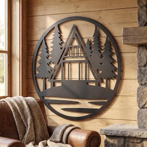 May include: A circular black metal wall art piece featuring a cutout design of an A-frame cabin nestled among several pine trees. The silhouette of the cabin includes a balcony and large windows, set against a backdrop of a forest landscape.