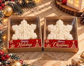 May include: Two white snowflake-shaped candles in brown cardboard boxes with clear plastic lids. The left box features a red label with gold text reading Happy Holidays, and the right box has a red label with gold text reading Merry Christmas.