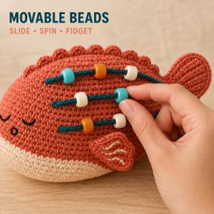 May include: A crocheted fish toy in rust orange and cream with three dark teal cords attached to its side. Each cord features movable beads in blue, orange, and white. The text MOVABLE BEADS SLIDE SPIN FIDGET is printed above the fish.