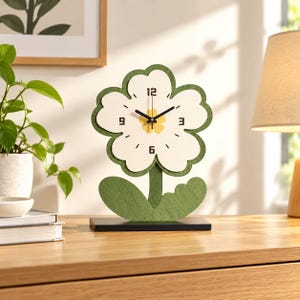 May include: A flower-shaped table clock with a white face, green border, and green stem base. The clock features black hands, black numerals at 3, 6, 9, and 12, and a yellow center detail. It stands on a rectangular black base.