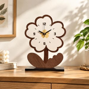 May include: A flower-shaped table clock with a dark brown wooden frame and a cream-colored face. The clock features black hour and minute hands, a yellow floral center, and black numerals at the 3, 6, 9, and 12 positions. It stands on a rectangular base.