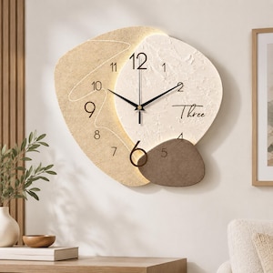 Puede incluir: Un reloj de pared abstracto con un diseño texturizado en capas de color beige y crema. La esfera presenta números negros, manecillas negras finas y la palabra Three escrita en cursiva. Una pieza de acento marrón oscuro se sitúa abajo con iluminación.