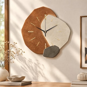 May include: A wall clock featuring an irregular, layered design composed of three distinct, textured shapes in shades of terracotta, cream, and dark gray. The clock face includes thin gold hour markers and black hands, with the numbers 12 and 6.
