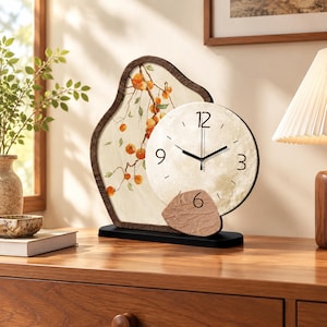 May include: A table clock featuring an irregular, dark-rimmed frame with a painted branch of orange fruit. A circular clock face with black hands and numerals 3, 6, 9, and 12 is mounted on the front, accented by a textured, stone-like piece.