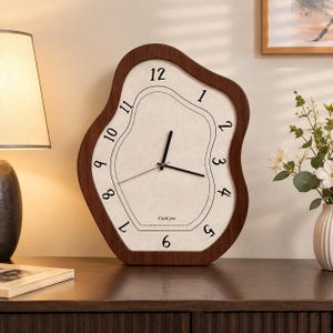 Peut inclure: Une horloge murale de forme irrégulière, semblable à une amibe, avec un cadre en bois brun foncé et un cadran crème. L'horloge présente des chiffres arabes noirs de 1 à 12, des aiguilles noires pour les heures et minutes, une trotteuse et le texte Card jue.