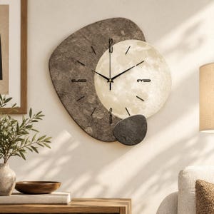 May include: A wall clock featuring an asymmetrical, textured dark gray base layered with a circular, cream-colored moon-like face. The clock has thin black hands and black numerical markers at the 12, 3, 6, and 9 positions, with a small dark gray accent piece.