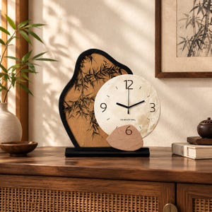 May include: A table clock featuring a circular cream-coloured face with black numerals 3, 6, 9, and 12, and the text UNI-BEAUTY MALL. The clock is set against an irregular black frame with a tan background displaying a black bamboo illustration.