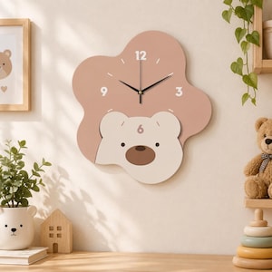 May include: A wall clock with an irregular, cloud-like shape in a dusty rose color. The clock face features a cream-colored bear head at the bottom, black clock hands, and white numerals 12, 3, 6, and 9 printed on the surface.