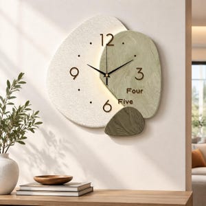 May include: A wall clock with an irregular, layered design featuring a large white textured section and a smaller sage green textured section. It has black hands, gold numerals 12, 3, 6, 9, and the words Four and Five printed in gold.
