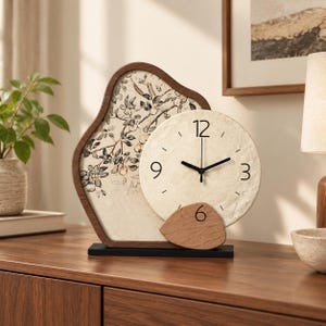 May include: A table clock with a dark wood frame featuring an irregular shape and a circular clock face. The face has black numerals 12, 3, 6, and 9. A decorative panel behind the clock displays a black and white botanical illustration.