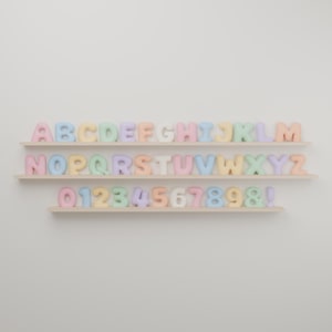 Puede incluir: Un conjunto de tres estantes de madera clara que muestran una colección de letras tridimensionales redondeadas en tonos pastel de la A a la Z, números del 0 al 9, un ampersand y un signo de exclamación en orden alfabético y numérico.