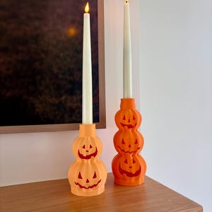 May include: Two orange pumpkin-shaped candle holders made of plastic, one featuring two stacked jack-o'-lantern faces and the other featuring three. Each holder contains a tall, slender, off-white taper candle with a glowing flame at the top.