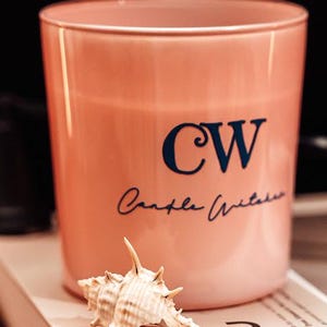 Puede incluir: Un recipiente cilíndrico de vidrio color melocotón con las letras azul oscuro CW y el texto en cursiva Candle Witches impreso en el frente. Una pequeña concha marina puntiaguda de color blanco roto se sitúa frente al recipiente.