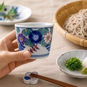 以下が含まれることがあります： 青、緑、赤の色合いで手描きされた鮮やかな花柄が特徴の白い陶器のカップ。カップは滑らかな仕上げの円錐形の円筒形で、内側は清潔な白で、複雑な植物のディテールが描かれています。