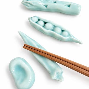 以下が含まれることがあります： アスパラガス、さやえんどう、細長い野菜、豆の形をした水色の陶器製箸置き4点セット。細長い野菜の形をした箸置きの上に、茶色の木製箸が1膳置かれています。