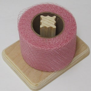 May include: A roll of pink tulle ribbon on a wooden spool, sitting on a wooden base.