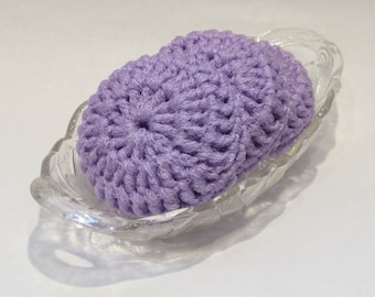 May include: A lavender crocheted soap dish sits in a clear, decorative glass dish. The soap dish is oval-shaped and has a textured, handmade appearance. The glass dish has a scalloped edge.