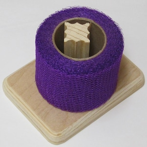 May include: A roll of vibrant purple mesh ribbon sits atop a light-colored wooden base. The ribbon is wrapped around a cardboard core, with a wooden support structure visible inside. The base has rounded corners.