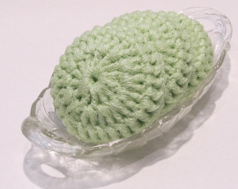 May include: A light green, crocheted soap saver sits in a clear, oval-shaped glass dish. The soap saver is a soft, textured oval shape. The dish has a decorative, scalloped edge and two small handles.
