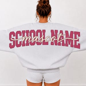 May include: A light grey crewneck sweatshirt with long sleeves and matching shorts. The back of the sweatshirt features the text SCHOOL NAME in large, bold, dark pink block letters with the word mascot in a cream-coloured cursive script overlay.