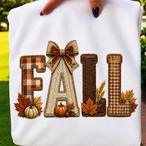 Op de afbeelding: Een wit sweatshirt met het woord FALL in grote, gestructureerde letters. De F heeft een bruin ruitpatroon, de A is crèmekleurig kabelbreisel met een strik, de L heeft een donkerbruine leertextuur en de tweede L heeft een bruin pied-de-poule patroon.