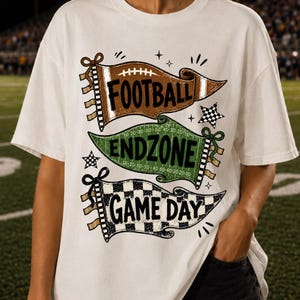 May include: A white t-shirt featuring a graphic design of three stacked pennants. The top brown pennant reads FOOTBALL, the middle green pennant reads ENDZONE, and the bottom black and white checkered pennant reads GAME DAY with small stars.