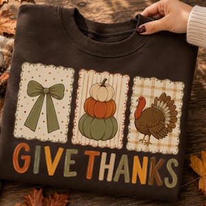 May include: A dark brown crewneck sweatshirt featuring three rectangular patches with scalloped edges. The patches display a green bow, a stack of three pumpkins, and a turkey. Below the graphics, the text GIVE THANKS is printed in large, multi-colored block letters.