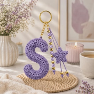 Puede incluir: Un llavero con la letra S tejida a ganchillo en color lavanda, con un anillo de metal dorado, una hilera de cuentas doradas y perlas lavanda, y un pequeño dije de estrella tejido a ganchillo. El accesorio tiene costuras detalladas.
