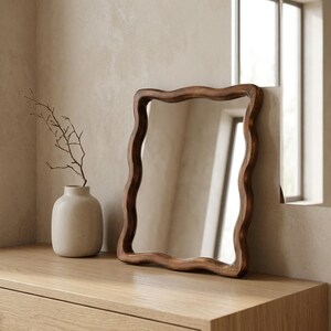 Small wavy wooden mirror with organic curved frame styled on a light oak console in a warm minimalist Japandi interior.