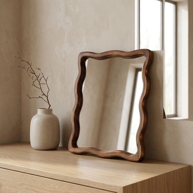 Small wavy wooden mirror with organic curved frame styled on a light oak console in a warm minimalist Japandi interior.