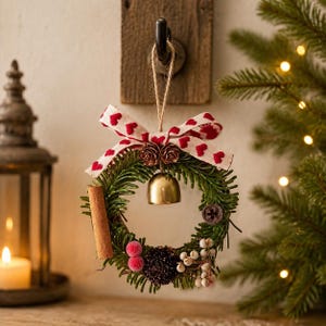Könnte beinhalten: Ein kleiner runder Kranzschmuck mit grünen Tannennadeln, einer zentralen Messingglocke, einer Zimtstange, roten Beeren, weißen Beeren und Tannenzapfen, verziert mit einer cremefarbenen Schleife mit rotem Herzmuster an der Oberseite.
