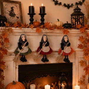 May include: Three hanging witch dolls with black headscarves, dark tops, and textured skirts in green, red, and orange. Each doll holds a small straw broomstick. The dolls feature pale faces and black shoes, suspended as decorative ornaments.