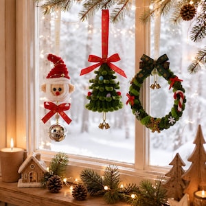 Op de afbeelding: Drie hangende kerstornamenten: een gehaakte kerstman met een rode muts en zilveren bel, een groene kerstboom van garen met een rode strik, en een groene krans met een donkergroene ruitjesstrik, kleine belletjes en een zuurstok.