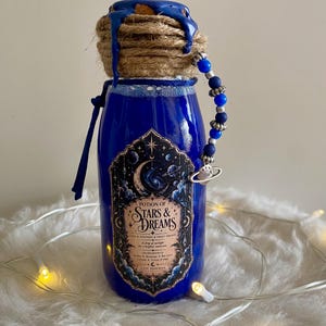 Peut inclure: Une bouteille en verre bleu étiquetée Potion of Stars & Dreams, dotée d'un bouchon en liège scellé à la cire bleue et entouré de ficelle de jute. Une breloque perlée aux accents bleus et argentés pend au col, orné d'un ruban bleu.