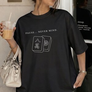 May include: A black short-sleeve t-shirt featuring a white graphic of two mahjong tiles and the text "PAUSE... NEVER MIND." The shirt is paired with light blue denim jeans and a beige handbag, while a clear plastic cup with a tan beverage is held.