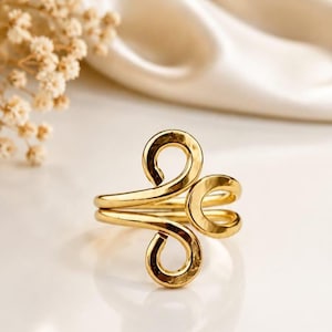 Puede incluir: Un anillo de tono dorado con una superficie pulida y reflectante de diseño abierto. La banda consta de dos bucles simétricos en forma de espiral que se extienden hacia afuera, creando una forma metálica fluida y continua.