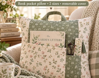 May include: A square reading pillow with a handle, featuring a floral pattern in green and pink. The pillow has a front pocket containing a book titled The Garden Letters, a pair of glasses, and a bookmark with a green tassel.
