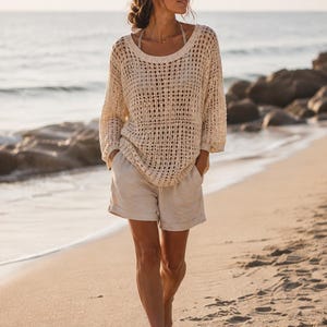 May include: A cream-colored, long-sleeved, open-knit sweater with a wide scoop neckline and a loose, relaxed fit, paired with light beige, solid-colored shorts featuring a cuffed hem and side pockets, creating a coordinated two-piece ensemble.