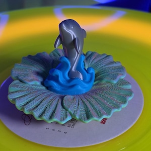 May include: A gray dolphin figurine emerging from blue waves, centered on a circular, textured, iridescent shell-like base with pink and green hues. The object rests on a white record label with black text, set against a bright yellow vinyl disc.