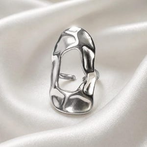 Pode incluir: Um anel de tom prateado com uma forma aberta, irregular e orgânica, com uma superfície lisa e reflexiva. A banda é dividida e ajustável, exibindo um design fluido semelhante a metal fundido que cria um centro oco na estrutura.