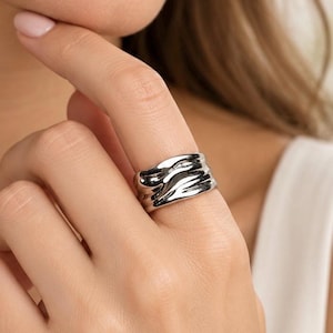 Könnte beinhalten: Ein breiter silberfarbener Ring mit einer polierten, reflektierenden Oberfläche, die unregelmäßige, wellenförmige und organische Erhebungen aufweist, welche sich um das gesamte Band ziehen und eine fließende Optik erzeugen.