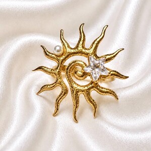 Pode incluir: Um anel de tom dourado com um design em forma de sol e superfície texturizada. O motivo solar inclui uma pequena pérola branca num dos raios e um cristal transparente facetado em forma de estrela posicionado perto do centro em espiral.