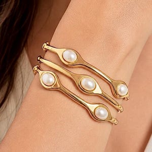 Pode incluir: Um conjunto de três pulseiras de metal em tom dourado, cada uma com um acabamento liso e polido e engastes ovais integrados. Cada pulseira é acentuada com pérolas brancas redondas e lustrosas incrustadas na estrutura de metal.