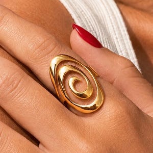Könnte beinhalten: Ein polierter Goldring mit einem großen, durchbrochenen ovalen Design und einem markanten zentralen Spiralelement. Die Metalloberfläche ist glatt und reflektierend und bildet ein durchgehendes Schleifenmuster, das den Finger mit einem Glanz überzieht.