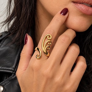 Op de afbeelding: Een goudkleurige open ring met een gepolijste afwerking. Het ontwerp bestaat uit twee afzonderlijke delen: de ene kant toont een enkele gebogen krul, terwijl de andere kant een groter, drielaags golfachtig patroon met spiraalvormige uiteinden heeft.