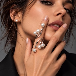 Könnte beinhalten: Ein silberfarbener Ring für mehrere Finger mit einem geschwungenen, durchbrochenen Design, das mit mehreren runden, weißen Kunstperlen verziert ist. Der Ring erstreckt sich über zwei Finger und hat zarte Metallstiele, die jeweils in einer Perle enden.