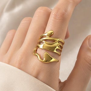 Könnte beinhalten: Ein goldfarbener Ring mit offenem, mehrschichtigem Design und fließenden, organischen, unregelmäßigen Formen. Das Band besteht aus mehreren miteinander verbundenen, glatten, polierten Metallbändern, die sich um den Finger legen.