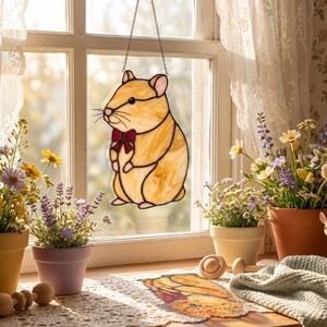 Peut inclure: Un attrape-soleil en vitrail en forme de hamster avec du verre beige clair, des yeux noirs et un petit nœud papillon rouge. Le hamster est suspendu par une fine chaîne métallique devant une fenêtre, avec une surface texturée et des lignes de plomb.